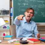 angry young male teacher wearing glasses pointing with index finger at camera shouting with aggressive expression sitting at school desk with books and notes in front of blackboard in classroom