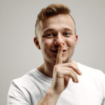 Keep silence. Handsome young man in white shirt looking at camera and holding finger on lips